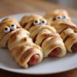 A plate of Halloween mummy hot dogs, baked and decorated with candy eyes.