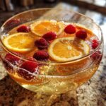 A close-up of a bubbly new years drink in a glass bowl, garnished with orange slices and fresh raspberries.