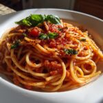 A close-up of a white bowl filled with spaghetti tossed in a rich tomato sauce, garnished with fresh basil and red pepper flakes. A perfect pasta dinner idea.