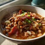 A steaming bowl of smoky crock pot black eyed peas recipe, garnished with bacon, onions, and green onions.