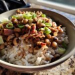 A close-up of a bowl filled with fluffy white rice topped with a hearty hoppin john recipe mixture of black-eyed peas, pulled pork, and green onions.