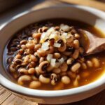 A close-up of a white bowl filled with Southern-style black eyed peas, topped with diced onions and a wooden spoon.