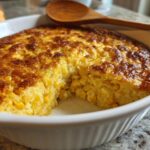 A close-up of a golden-brown corn pudding recipe baked in a white dish, with a serving spoon resting on top.