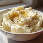 A bowl of creamy mashed potatoes recipe, topped with melted butter and cracked black pepper.
