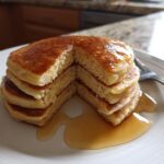 A stack of fluffy pancakes recipes drizzled with maple syrup on a white plate, with a fork to the side.