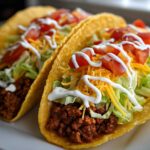 Close-up of two ground beef tacos filled with lettuce, cheese, tomatoes, and sour cream.
