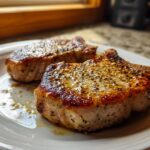 Two perfectly seared pork chops seasoned with pepper and herbs on a white plate, a delicious part of pork chops recipes.