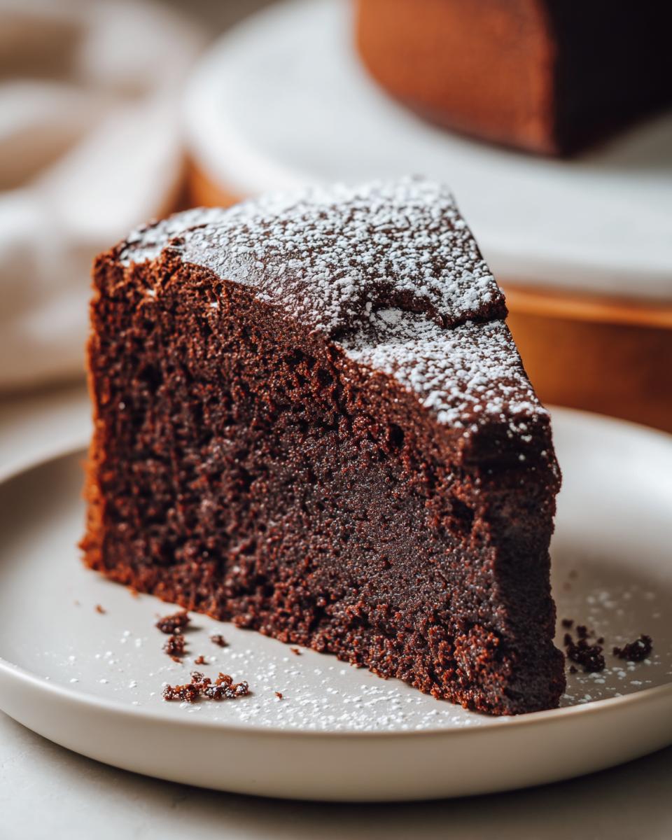 A moist slice of chocolate cake dusted with powdered sugar on a white plate