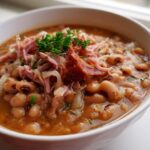A close-up of a white bowl filled with hearty black-eyed peas, shredded ham, and crispy bacon, a perfect new years day food.