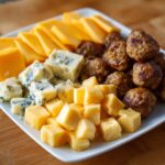 A white platter filled with various new year's eve snacks including cubed cheddar cheese, blue cheese crumbles, sliced cheddar, and mini meatballs.