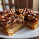 Close-up of three rich and gooey pecan pie bars on a white plate, showcasing the flaky crust and pecan topping.