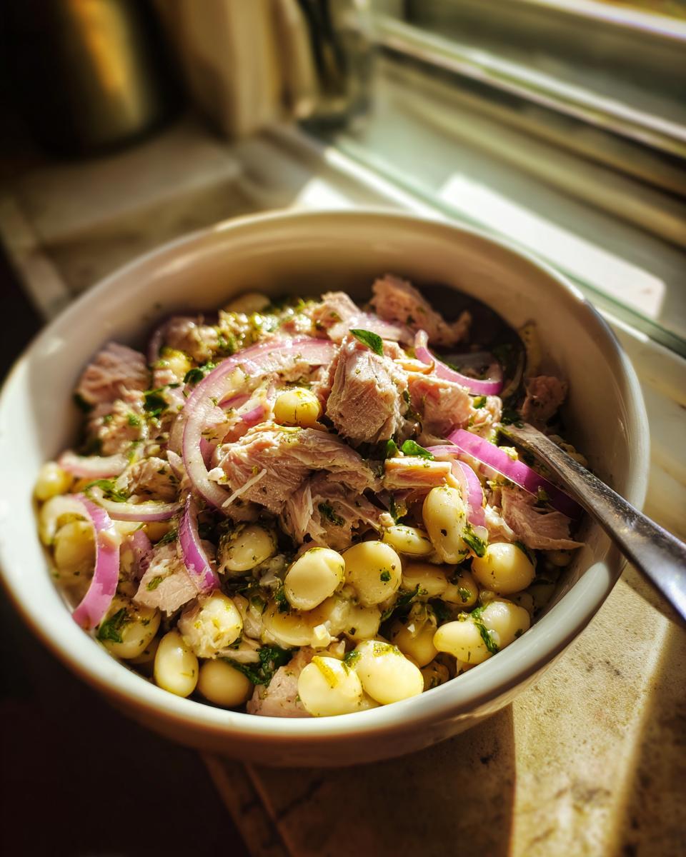 Bowl of tuna and white bean salad with lemon dressing, red onion slices, and herbs.