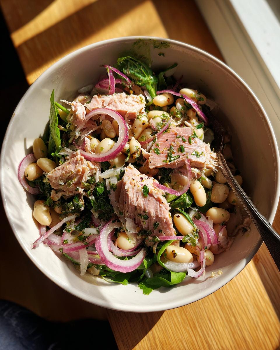 Bowl of tuna and white bean salad with lemon dressing showing tuna chunks, white beans, red onions, and greens