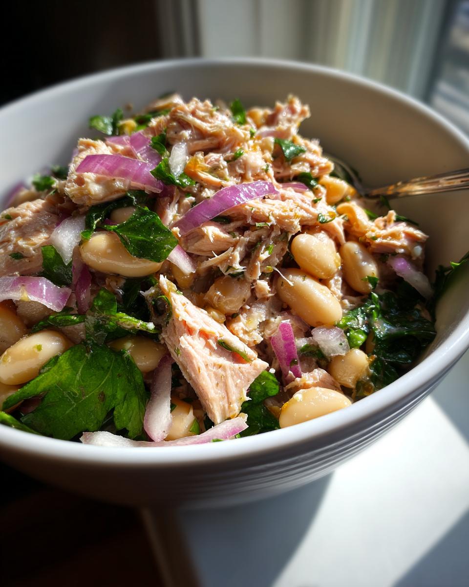 Close-up of tuna and white bean salad with lemon dressing, red onions, and fresh greens in a white bowl.