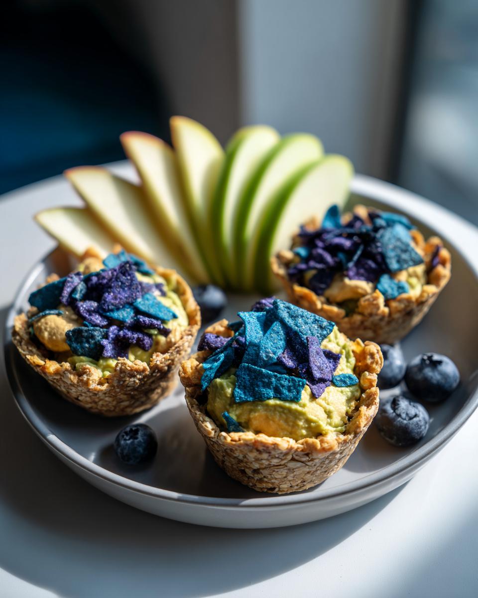 Three oat cups filled with green dip and blue chips, served with apple slices and blueberries on a plate.