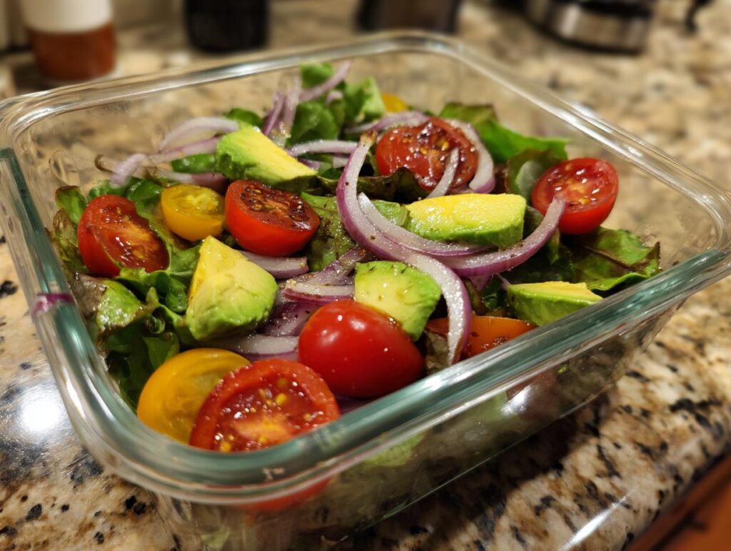 Glass container with a healthy packed lunch salad of avocado, cherry tomatoes, red onion, and leafy greens.